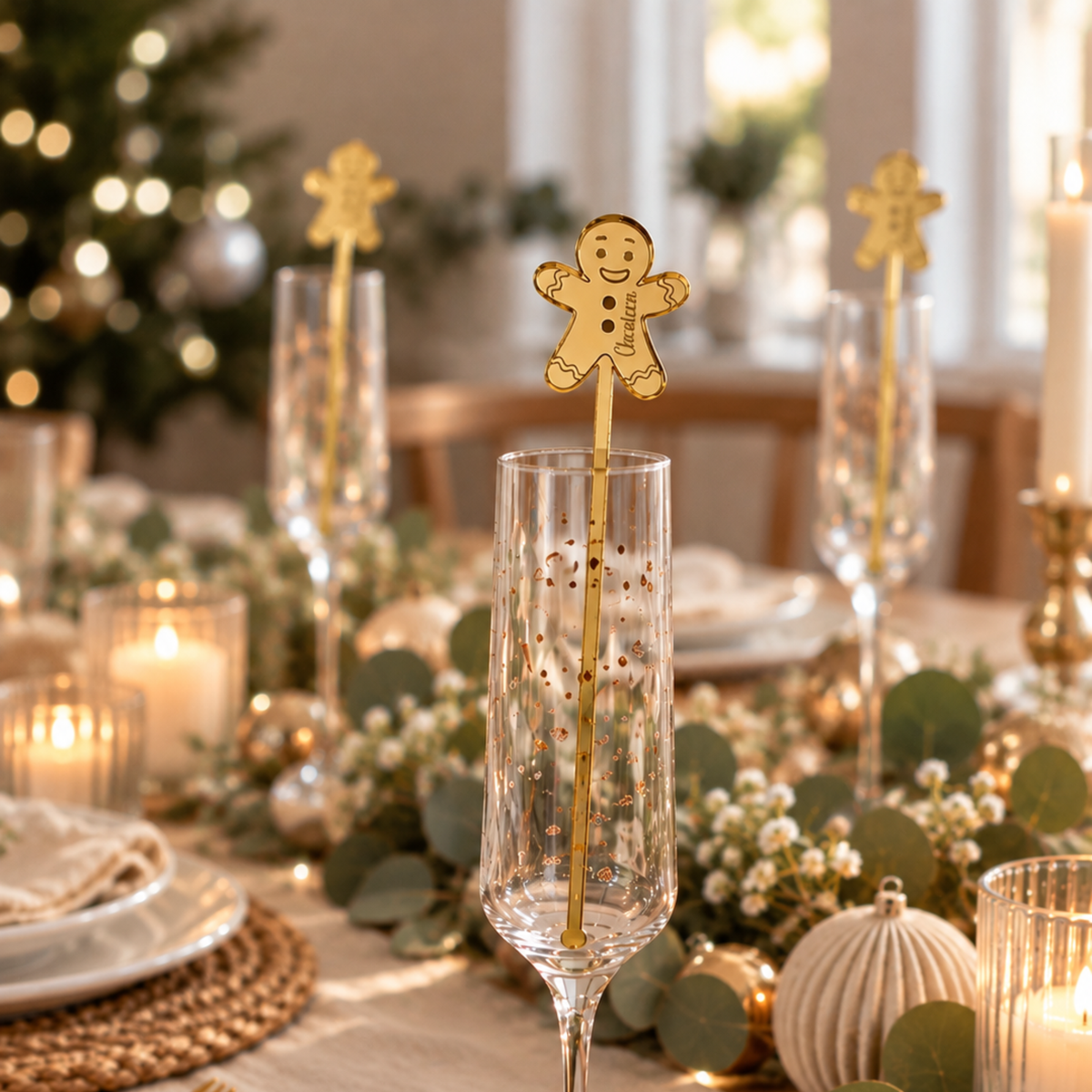 Personalised gold mirror acrylic gingerbread Christmas drink stirrer in a champagne flute, styled on an elegant festive table with matching stirrers, eucalyptus greenery and candles.